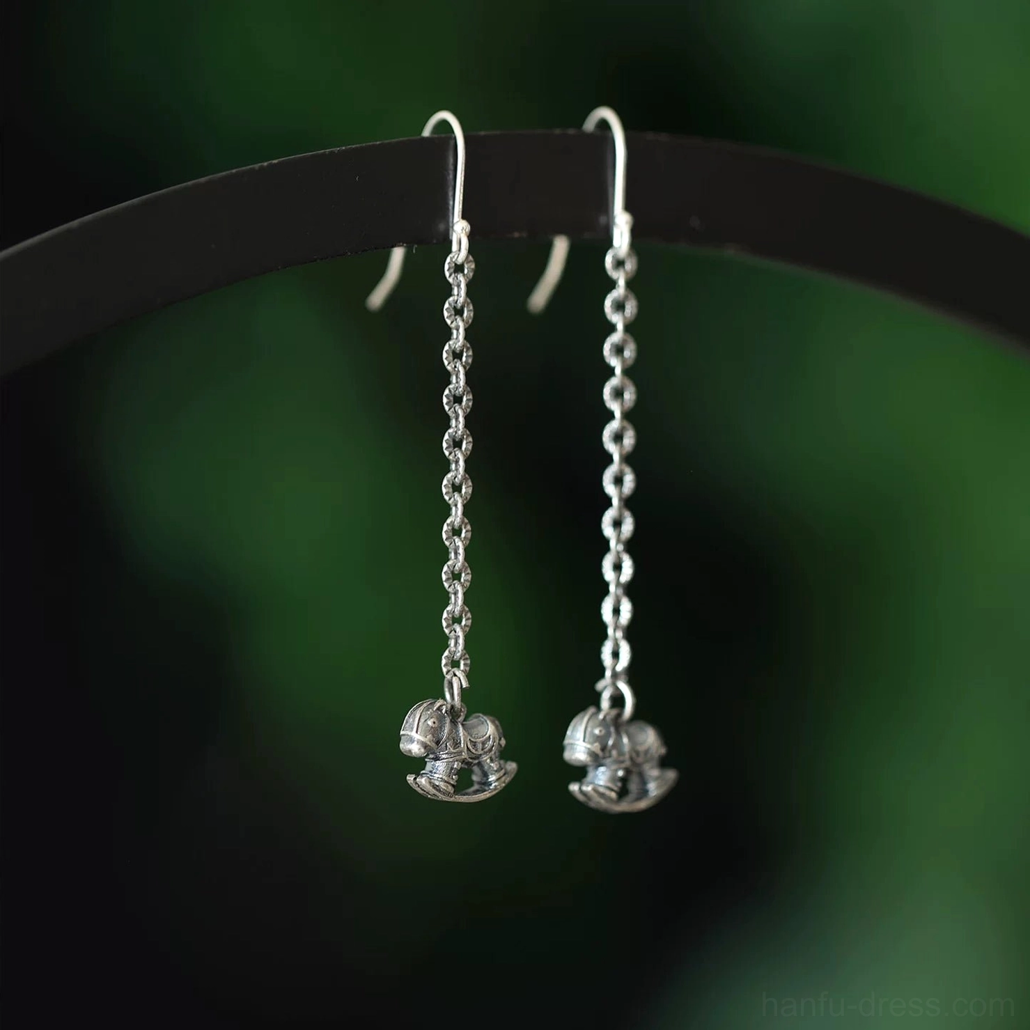 Rocking horse drop earrings with twisted cable chains and polished silver-toned 3D charms displayed on a black jewelry rod against blurred green foliage