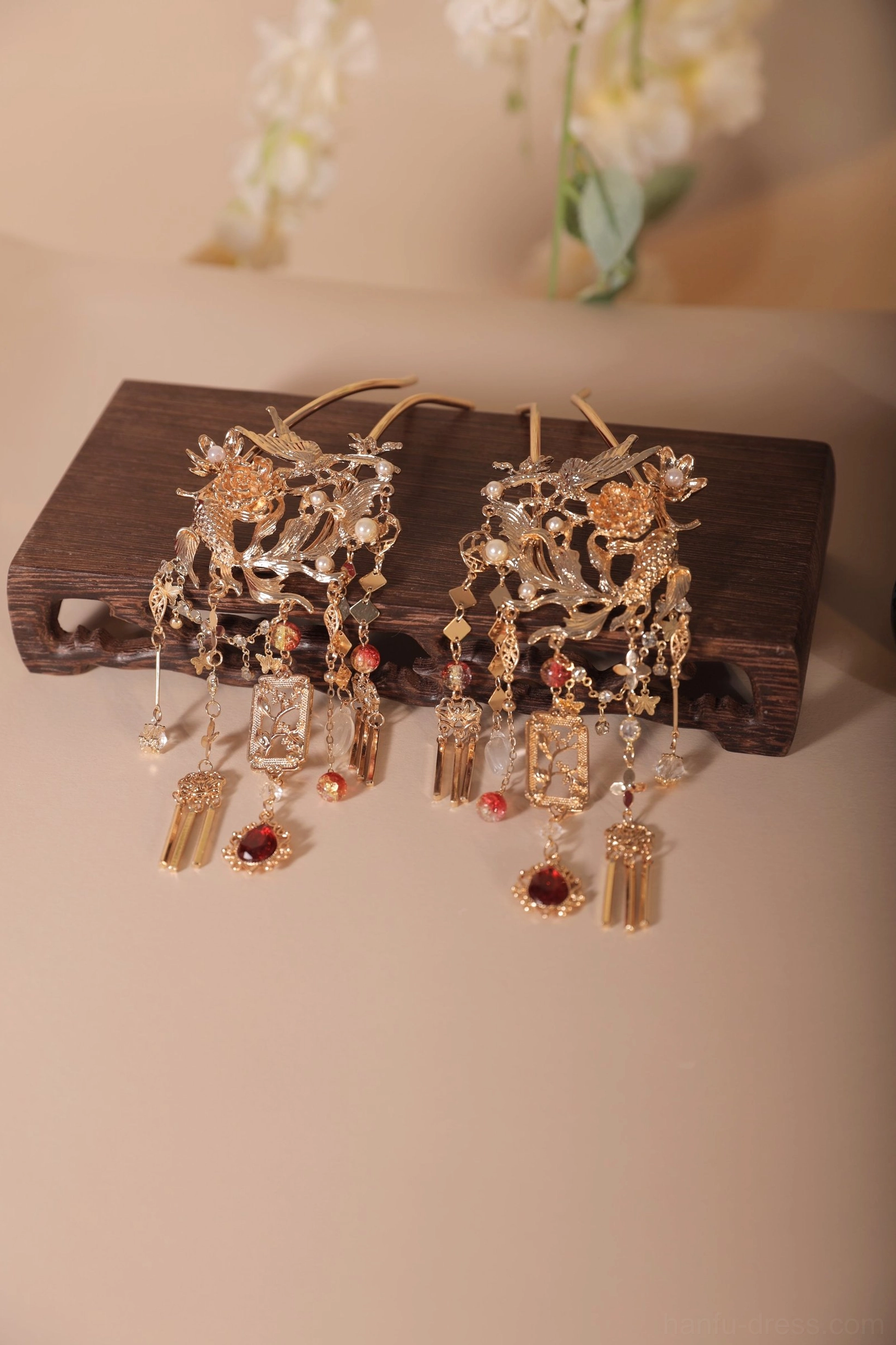 Pair of antique-inspired Chinese hair pins displayed on a dark wooden stand, with intricate gold-toned carving, red burgundy gemstone details, and delicate metal tassel components