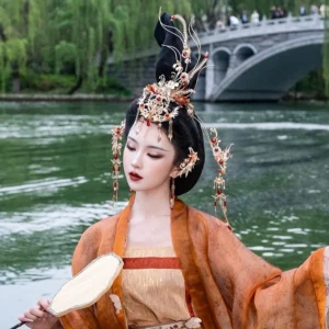 Ao Feng Yin phoenix Hanfu bridal crown and tassel hair accessory set worn with orange Tang style Hanfu beside a lakeside garden