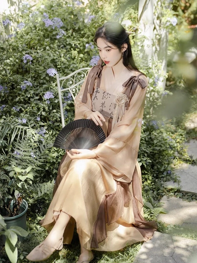 Apricot and brown modern Tang style Hanfu dress set with calligraphy print, sheer sleeves and black folding fan in a flower garden