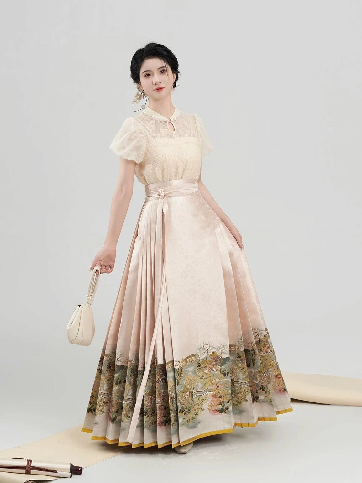 Side view of an apricot Chinese hanfu blouse and mamian skirt with gold trimmed landscape hem styled with a mini beige handbag