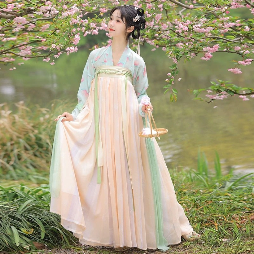 Three-quarter side view of an apricot gradient Tang Dynasty Hanfu Qixiong Ruqun with a soft flowing pleated skirt photographed beside a lakeside woodland