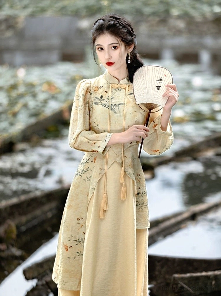 Front view of an apricot beige modern Hanfu cheongsam midi dress with ink floral print, stand collar, puff sleeves, front tassel pendants, and asymmetrical layered hem worn beside a Chinese lotus pond