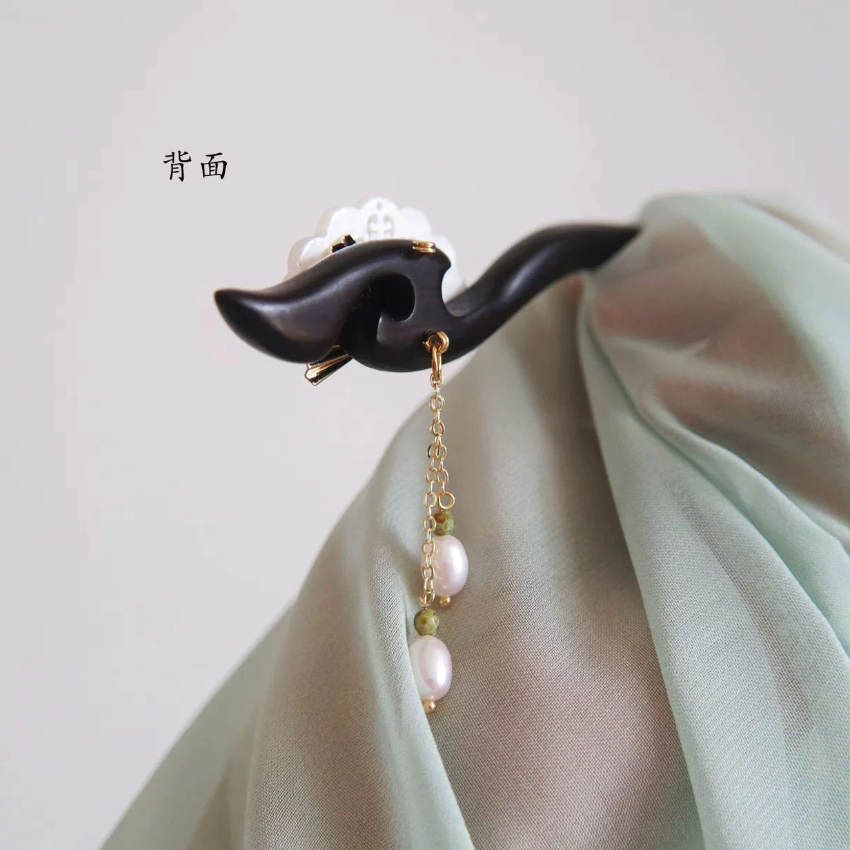 Back view of a curved black wooden Chinese hairpin with hidden tassel attachment, floral top ornament, and pearl drop detail on Hanfu fabric
