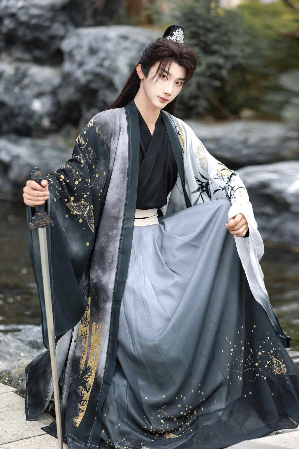 White men’s wuxia style hanfu full set in a rocky garden setting, showing wide sleeves, grey ink-wash gradient skirt, and gold foil bamboo details
