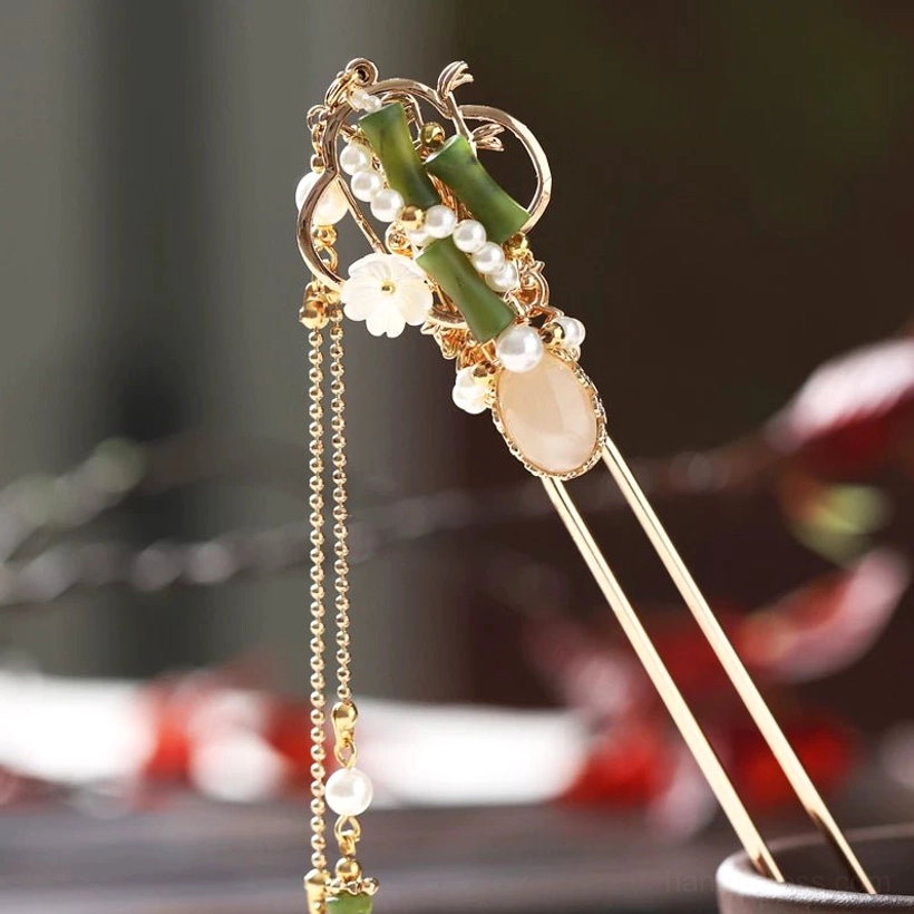 Front-right close-up of a bamboo-style tassel hair stick with gold-tone structure, green bamboo details, white flower charms, faux pearls, warm beige gem accent, and long pendant tassel
