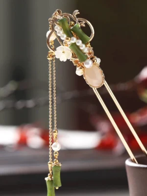 Close-up of a bamboo-inspired Chinese hair stick with green bamboo ornaments, white flower charms, faux pearls, warm beige centerpiece, gold-tone frame, and long hanging tassel on a dark wooden table