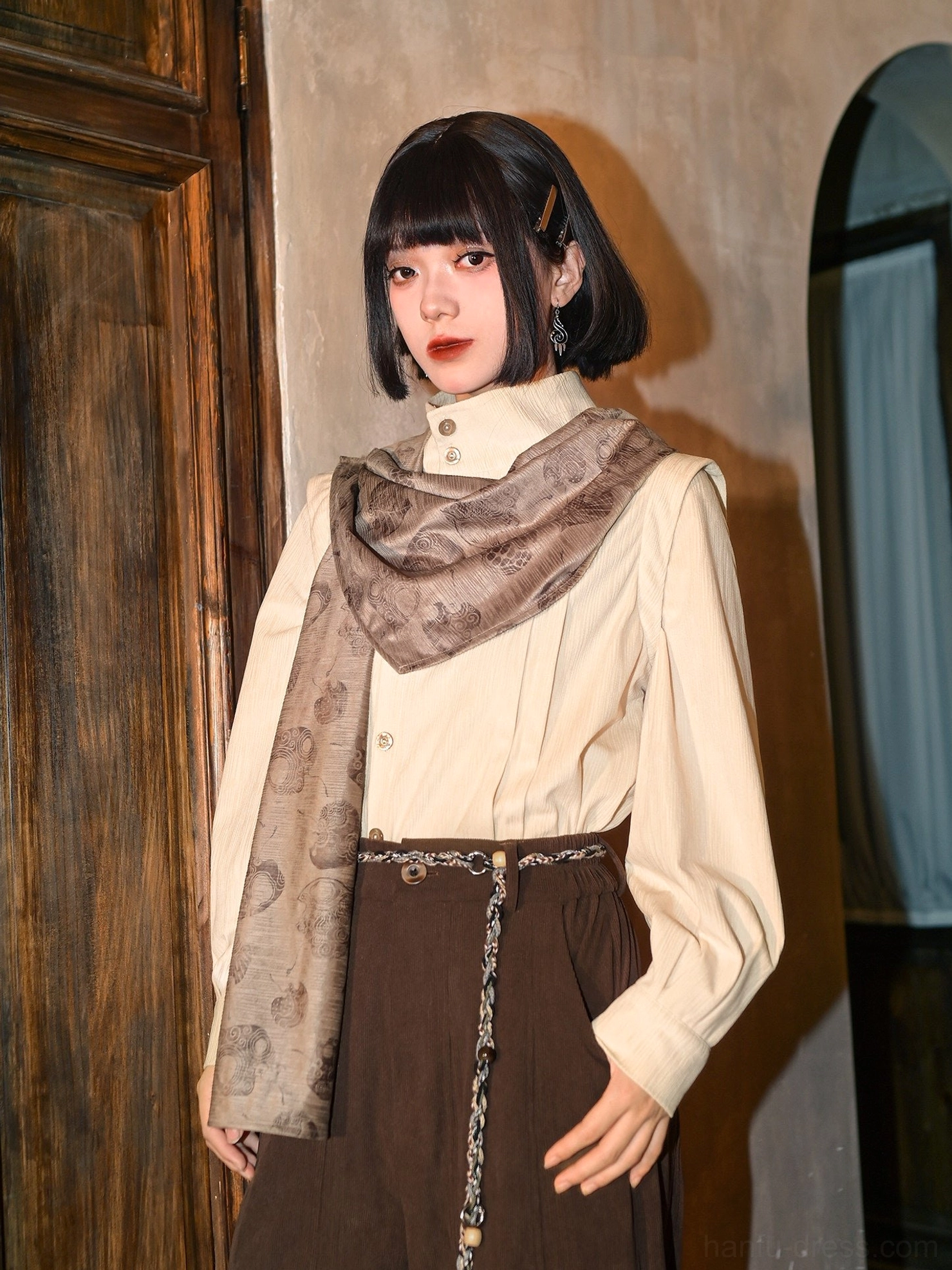 Three quarter front view of a beige modern Hanfu long sleeve blouse styled with scarf-like detachable cape, dark brown high waisted skirt, and tassel waist chain