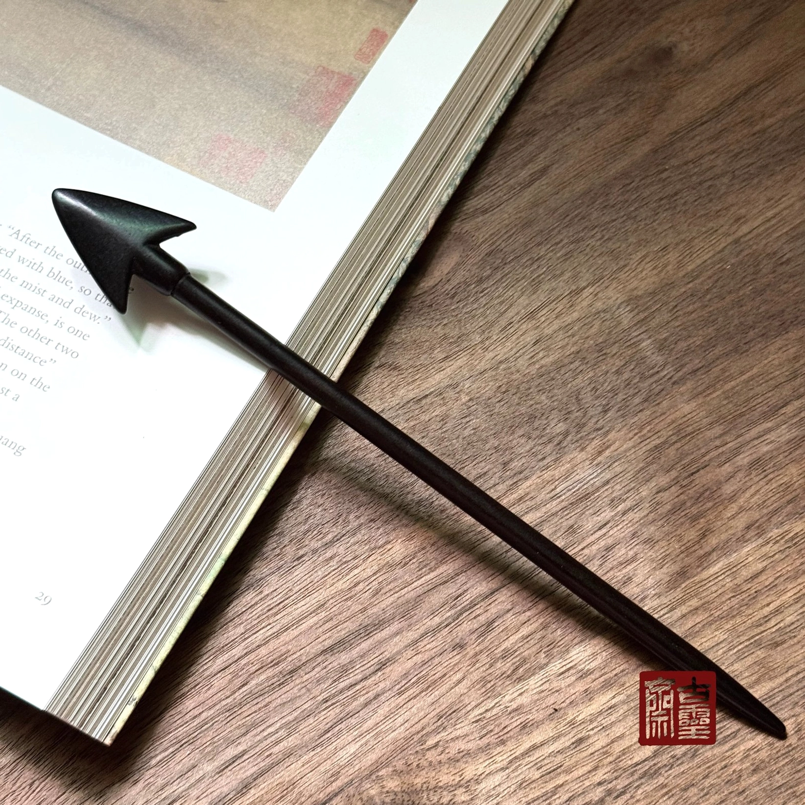 Matte black arrowhead Chinese wooden hair stick displayed on an open book and wood grain table for Hanfu hair styling