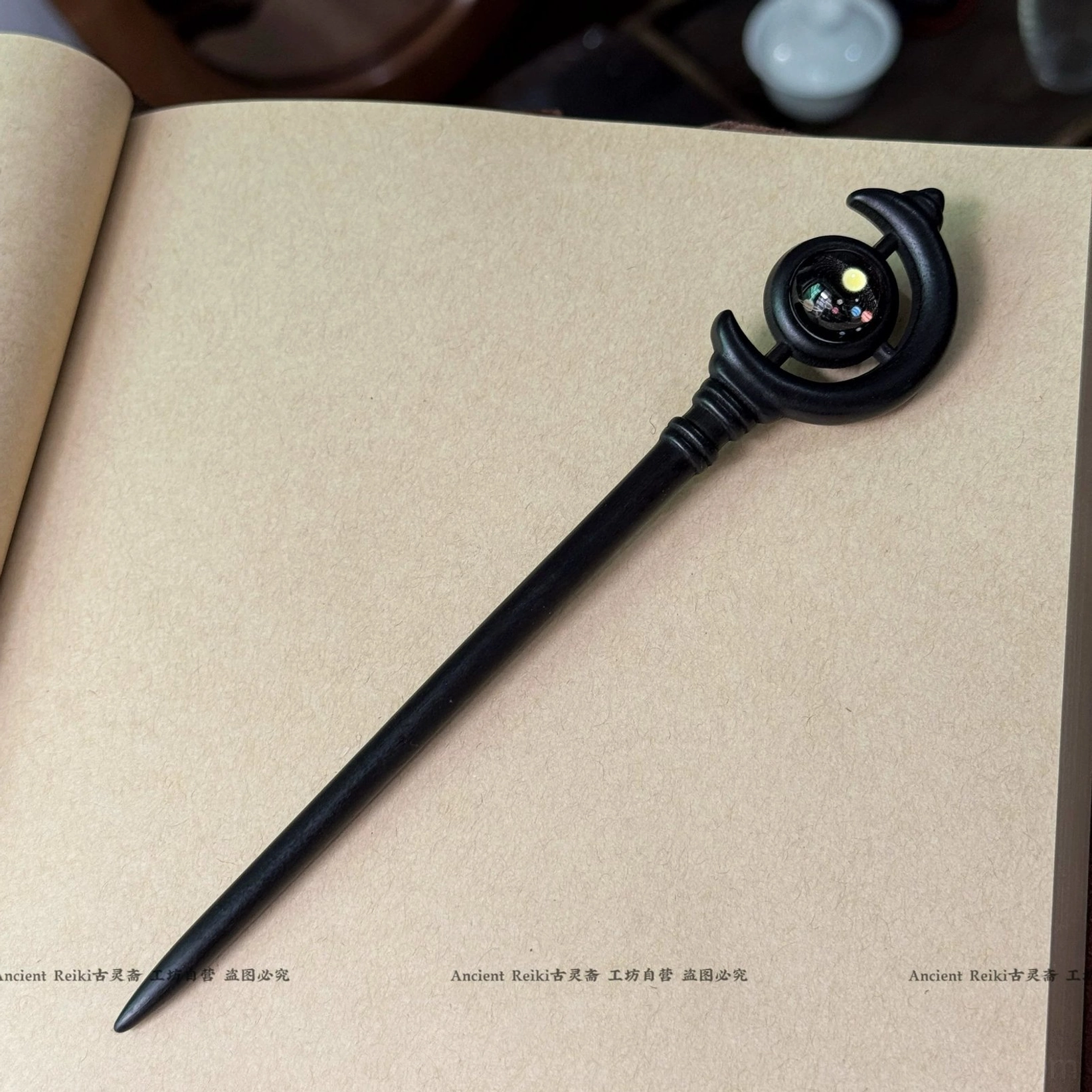Matte black Chinese hair stick with crescent moon top and colored glass bead displayed on an open kraft paper notebook beside traditional tea accessories