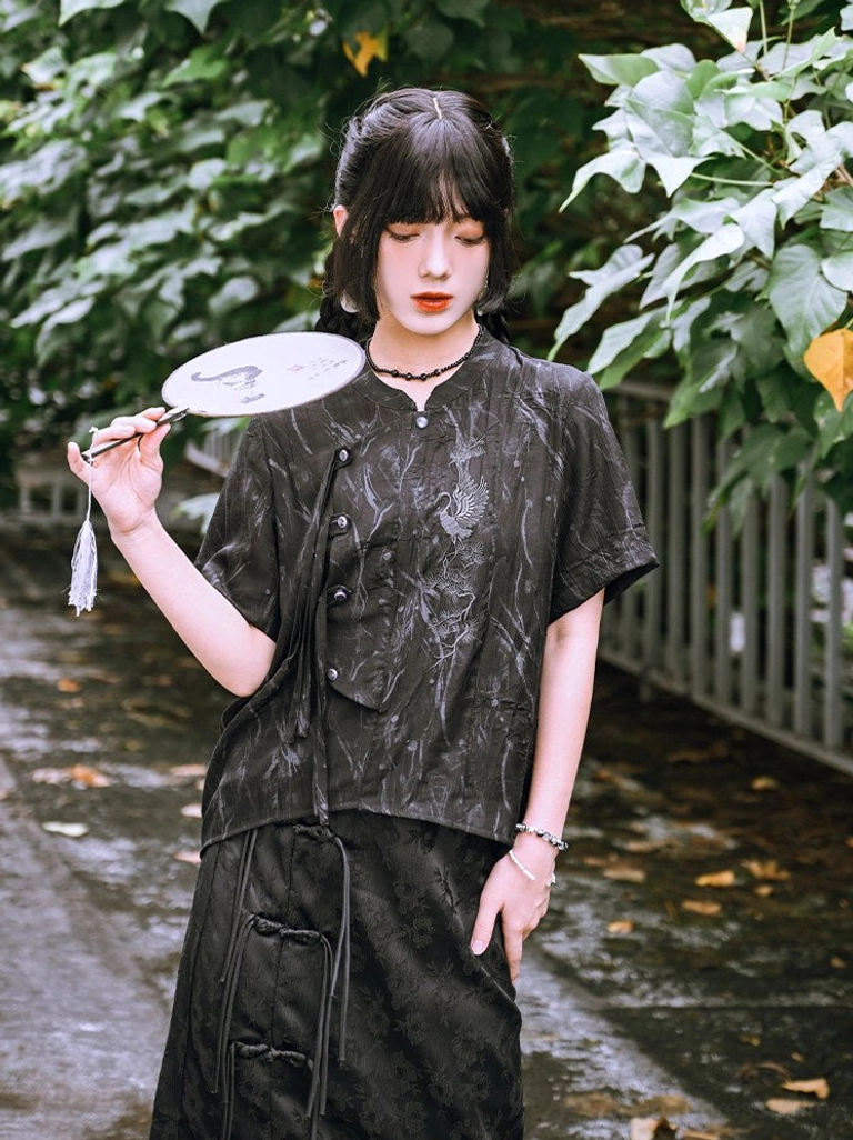 Black modern Hanfu short sleeve shirt with embroidered crane, asymmetrical frog buttons, stand collar, and tonal jacquard fabric styled with a matching skirt outdoors