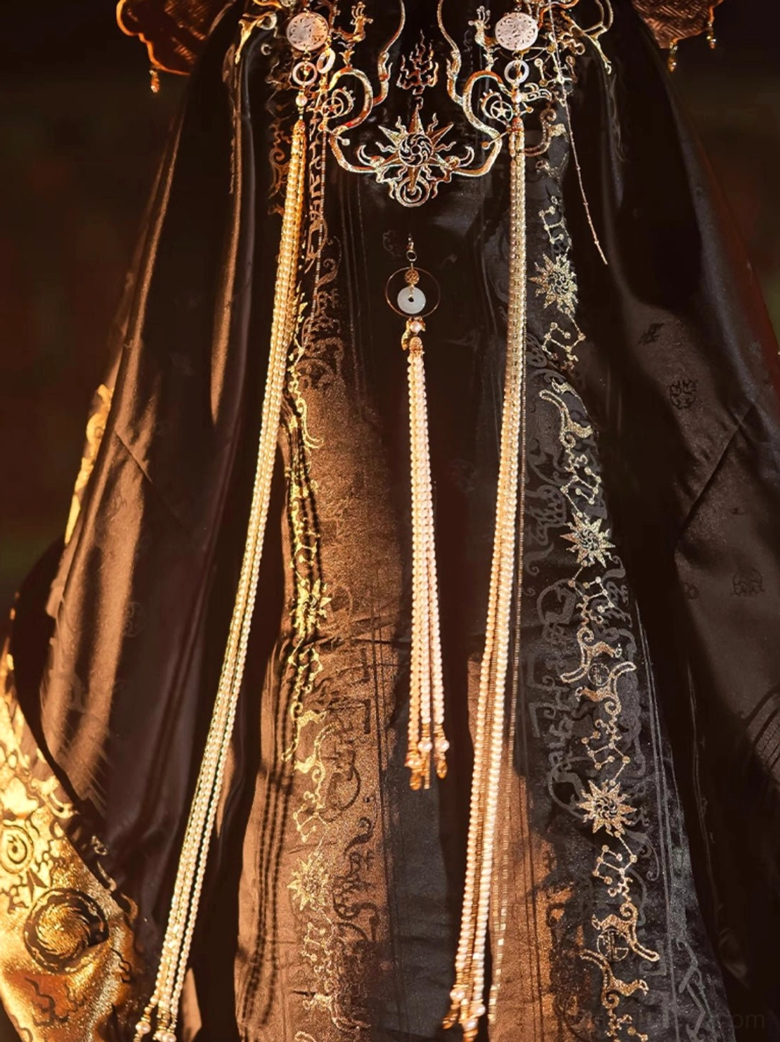 Close up of the black and gold fantasy hanfu accessory details showing an ornate royal metal ornament, long golden bead tassels, white jade style pendant, and traditional embroidered fabric