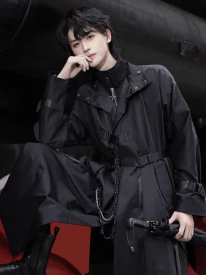 Three-quarter view of a black gothic Hanfu robe coat showing matte polyester fabric, decorative waist chain, snap-button stand collar, buckle-adjusted cuffs, and long tailored silhouette beside a red steam locomotive wheel