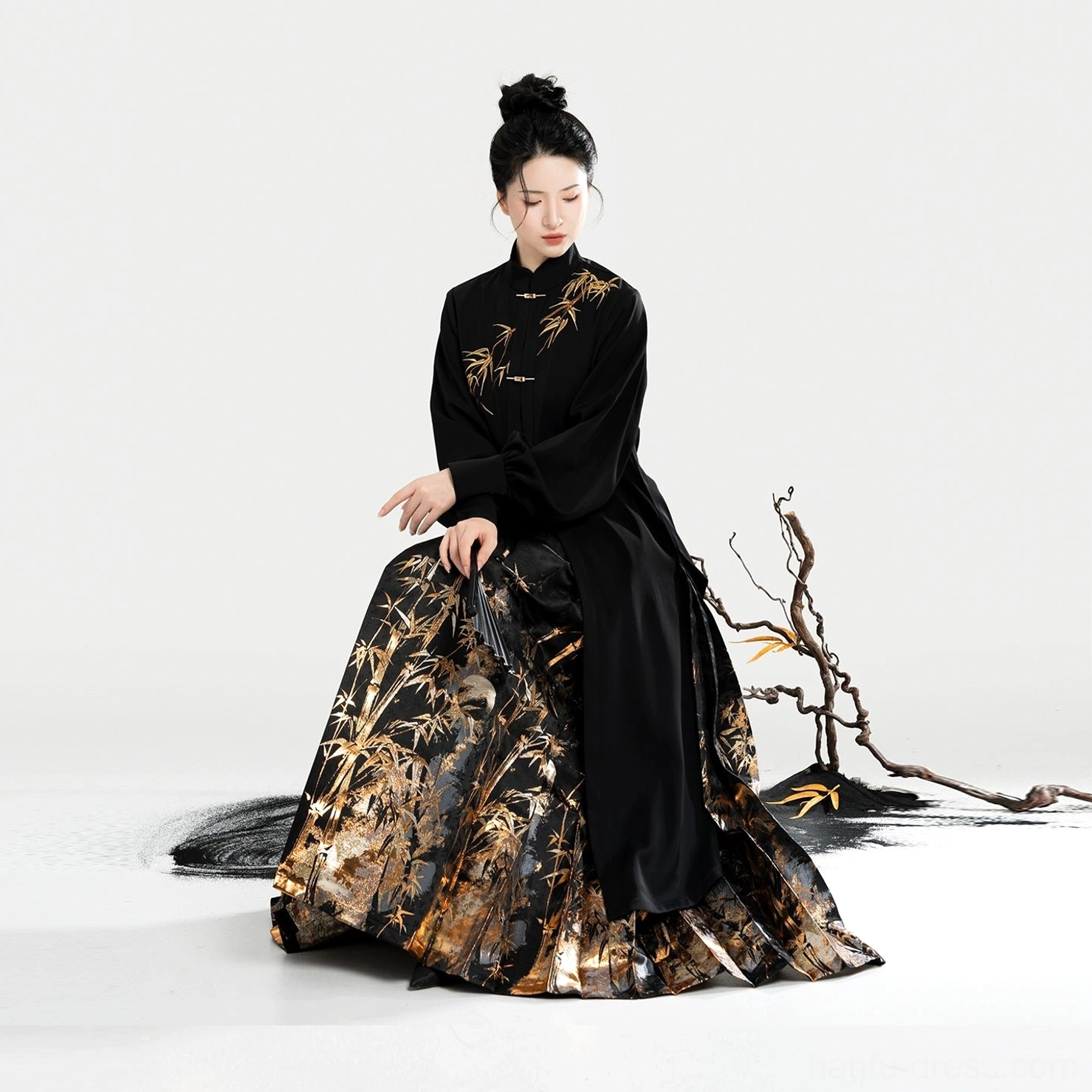 Seated three-quarter view of a black Hanfu blouse and golden bamboo mamian skirt with a folding fan