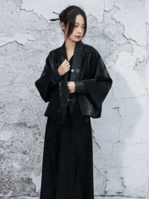 Black modern hanfu cropped outerwear coat shown in a front three-quarter view, featuring a ribbed collar, metallic buckle fasteners, relaxed boxy fit, and layered cuffs over a flowing black dress