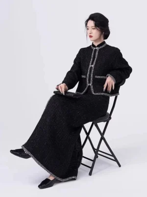 Seated three-quarter view of a black modern Hanfu outfit with tailored jacket, silver pocket trim, long wrap skirt, and black flats