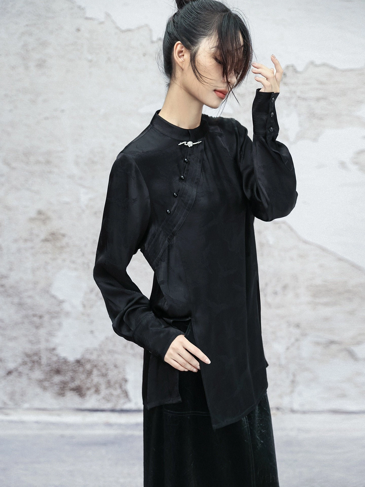 Side-front view of a black modern Hanfu satin top showing the asymmetric side hem, side slit, decorative frog button placket, and long sleeves