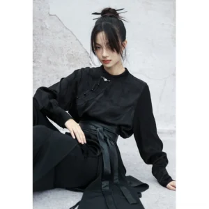 Front view of a black satin modern Hanfu long sleeve top with stand collar, frog buttons, subtle dark woven pattern, and relaxed fit styled with wide-leg trousers