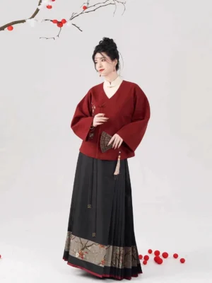 Front full-body view of a Blessing Branch Ming Dynasty hanfu set with a burgundy embroidered coat, black pleated mamian skirt, tassel fan, and festive red berry branch backdrop