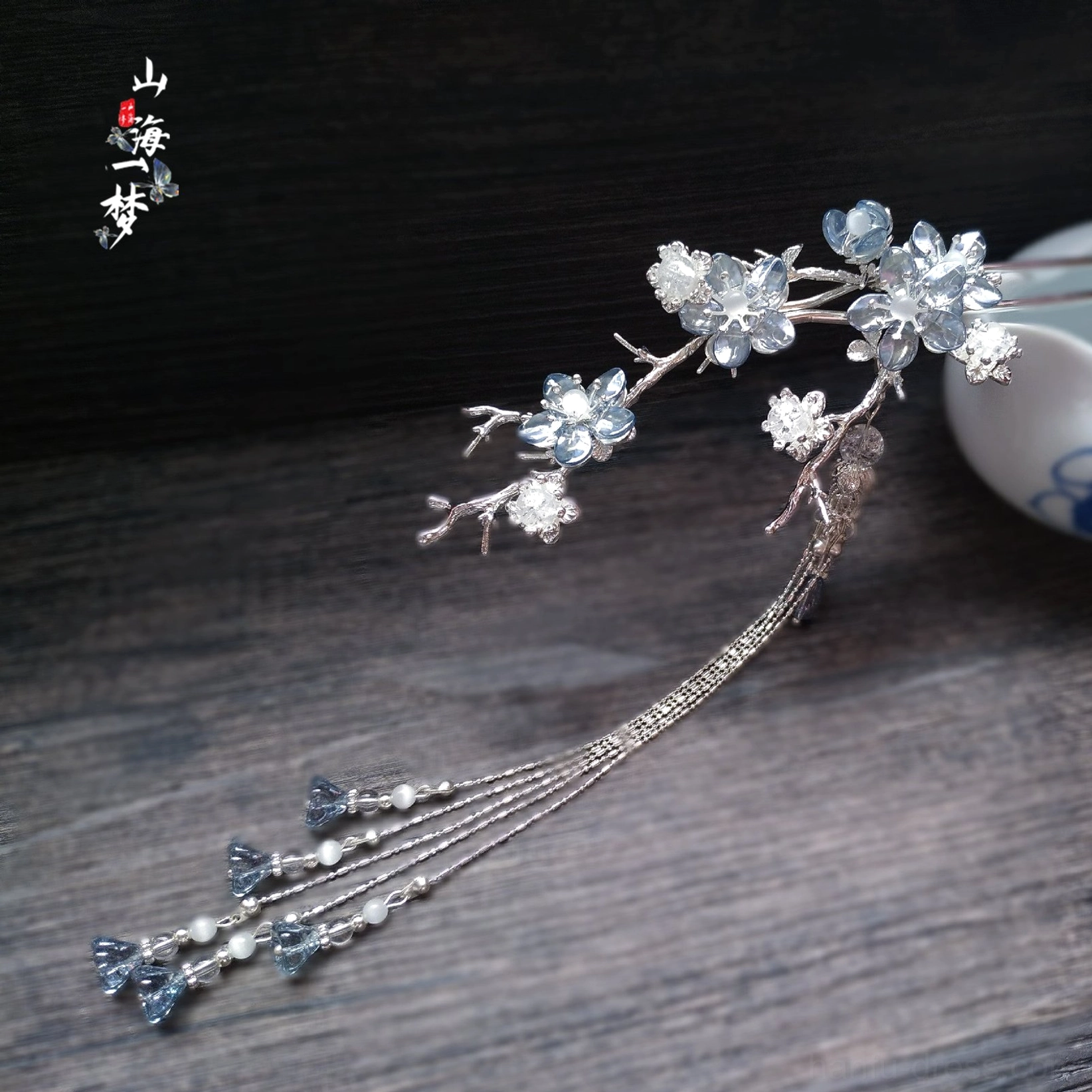 Blue crystal plum blossom hanfu hairpin with silver-tone branches and long layered tassels displayed on a dark wooden table
