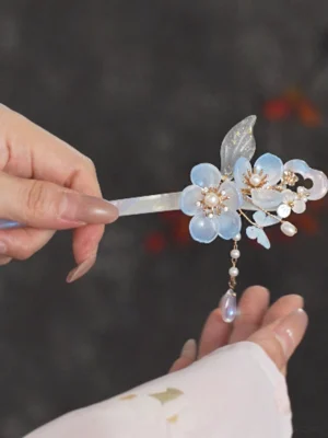 Hand holding a blue flower Chinese hair stick with translucent opalescent shaft and dangling blue tassel, styled with a pink Hanfu sleeve