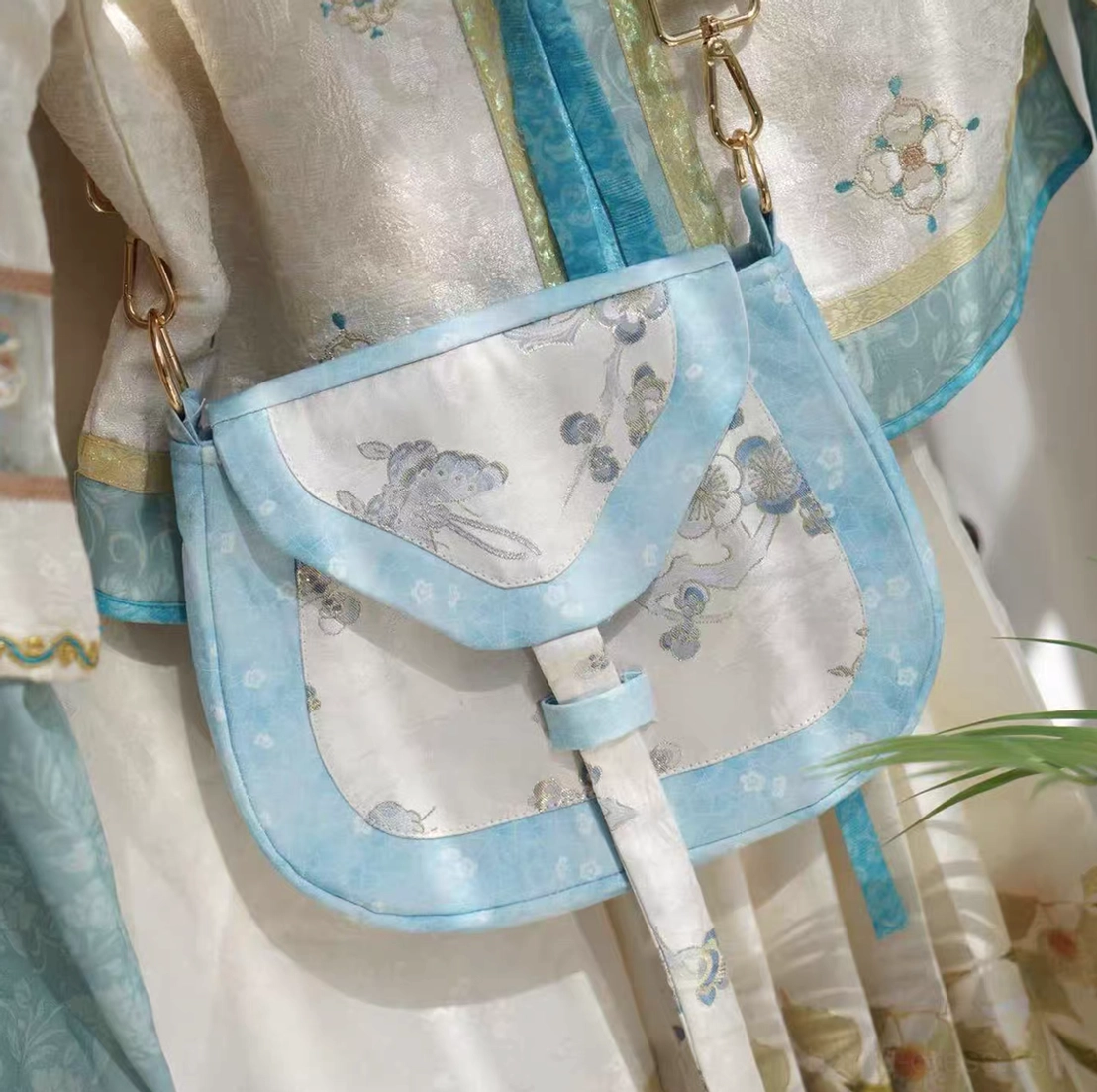 Three-quarter view of a traditional Chinese crossbody saddle bag with blue flower and rabbit embroidery, sky-blue trim, front strap closure, and gold-tone shoulder strap hardware