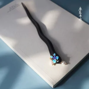 Blue flower Chinese hair stick flat lay on a notebook featuring a dark wavy wooden shaft and delicate pearl charms