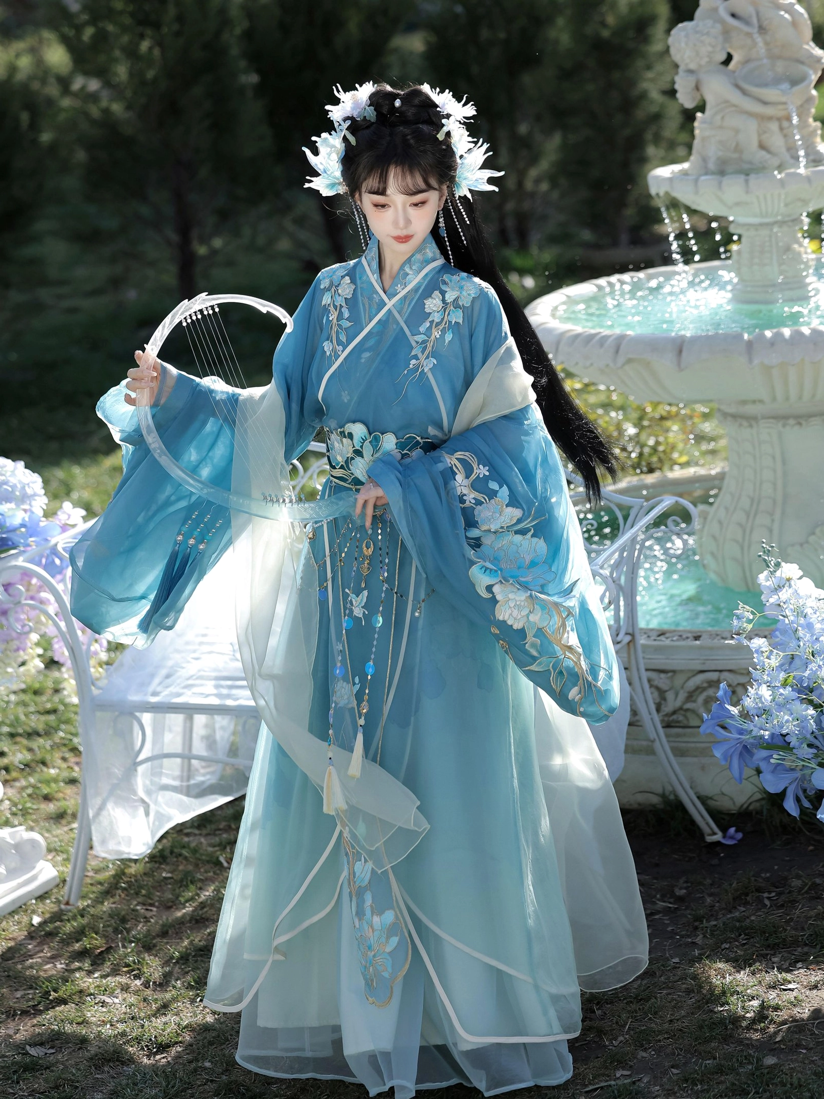 Elegant blue gradient traditional Hanfu dress with floral sleeve embroidery, decorative waist chain, and hanging tassel pendants in an outdoor garden setting