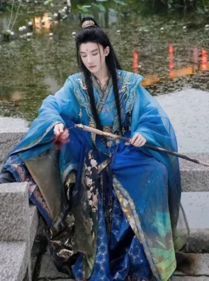Male model wearing blue gradient xianxia hanfu costume with wide sleeves beside a lotus pond at night