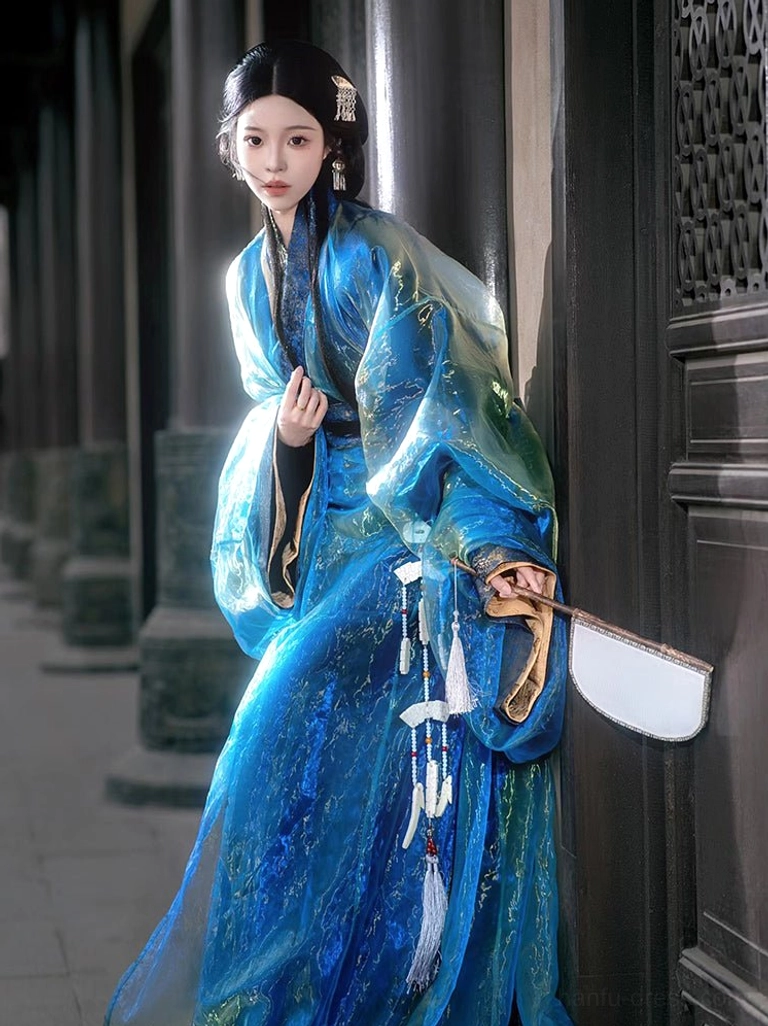 Side view of a blue-green shimmering Warring States and Han Dynasty Hanfu robe with voluminous wide sleeves worn with a flat white silk fan in an ancient courtyard
