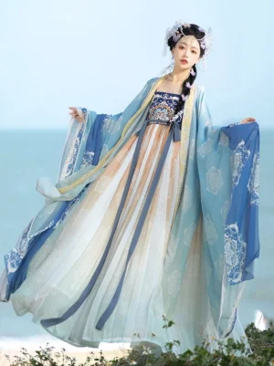 Blue and ivory gradient Tang Dynasty Hanfu Heziqun dress with embroidered chest panel, floral wide sleeve outer robe, and flowing silk-style waist sashes photographed beside the seaside