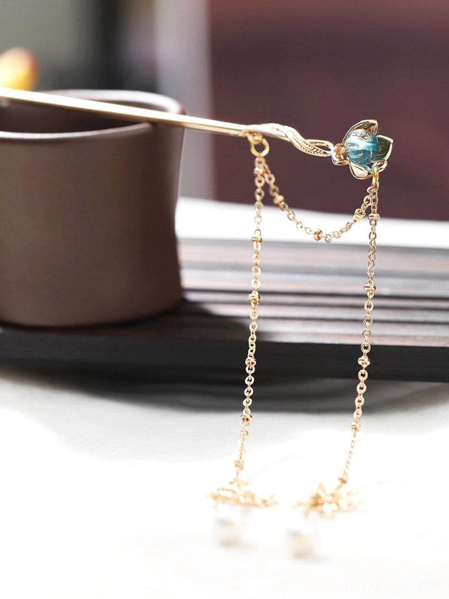 Blue lotus tassel hair stick for Hanfu leaning beside a cup, showing the gold-tone chains, pearl pendants, and decorative snowflake charms
