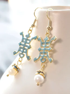 Light blue Shou longevity pearl drop Chinese earrings with gold tone hook closures displayed on a white ceramic vase