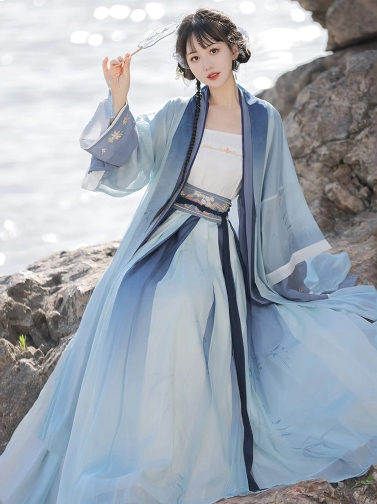 Three-quarter seated view of a blue gradient Song Dynasty Qiyao Ruqun Hanfu with pleated skirt, matching beizi robe, and delicate traditional hairpin by the seaside
