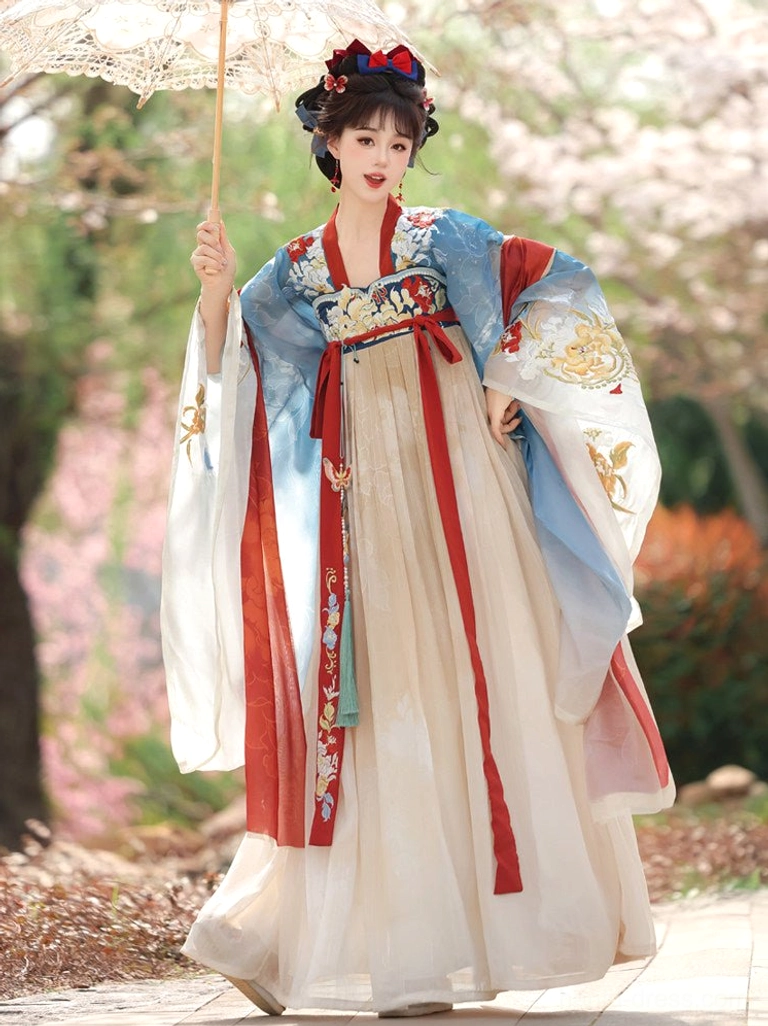 Front full-body view of a blue and cream Tang Dynasty Qixiong Ruqun Hanfu outfit with embroidered wide sleeves, red sashes, pleated skirt, and traditional hair ornaments in a spring flower garden