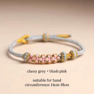 Blush pink and grey handwoven Chinese knot bracelet with floral woven charms, gold and silver tone decorative knots, and adjustable sliding knot closure on a light beige background