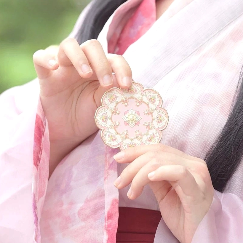 Woman wearing pink Hanfu holding a blushing lotus blossom Chinese hand mirror, showing the ornate embossed floral back and rose gold-tone petal edge