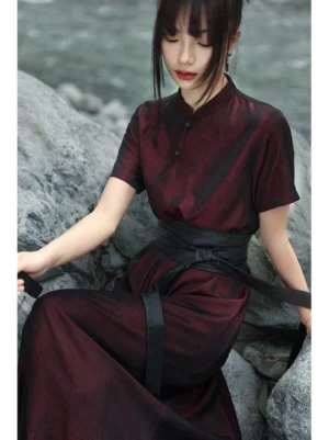 Close-up view of a burgundy Chinese cheongsam dress with stand collar, traditional frog fasteners, textured polyester fabric, and black waist sash styled on a seated model