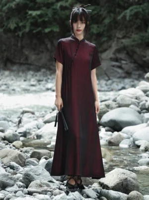 Full-body front view of a woman wearing a burgundy modern Hanfu cheongsam dress with black gradient hem, stand collar, short sleeves, diagonal frog fasteners, and flowing floor-length silhouette beside a rocky stream