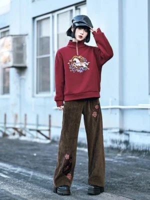 Full-length front view of a woman wearing a burgundy red modern Hanfu fleece sweatshirt with Chinese horse and cloud embroidery, stand collar, quarter-zip closure, brown trousers, and retro newsboy cap