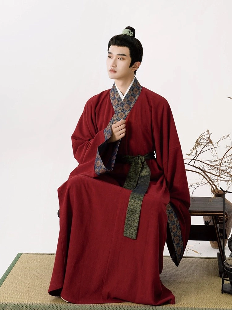 Seated male model wearing the burgundy red cross-collar inner robe from a Song Dynasty-inspired hanfu outfit with patterned collar trim and dark green waist sash