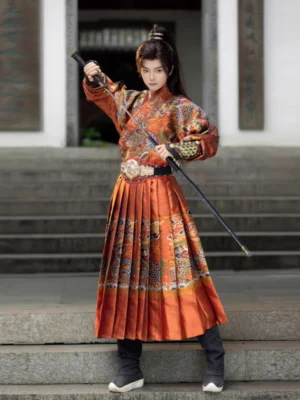 Burnt orange Ming Dynasty Jinyiwei Feiyufu hanfu for men shown from the front three-quarter view in a traditional Chinese courtyard, featuring ornate brocade patterns, a flowing robe, pleated lower garment, and decorative waist belt