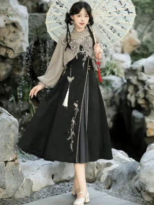 Full-body front view of a woman wearing the Butterfly Ash modern Hanfu summer sundress two-piece set with grey lantern-sleeve cropped top, black embroidered midi dress, butterfly cloud collar, and tassel pendant in a traditional Chinese garden
