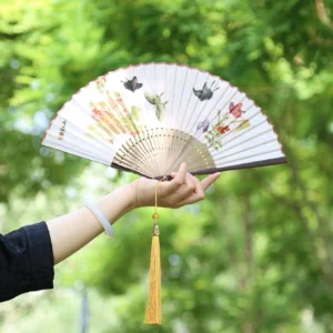 Butterfly floral traditional Chinese folding hand fan with off-white fan surface, dark brown bamboo ribs, Chinese calligraphy details, and golden-yellow tassel held outdoors against lush green foliage