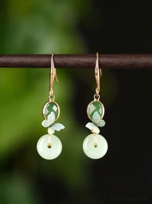 Butterfly jade drop earrings with gold hooks, green oval accents, enamel butterfly charms, and light green peace buckle pendants displayed against a dark botanical background