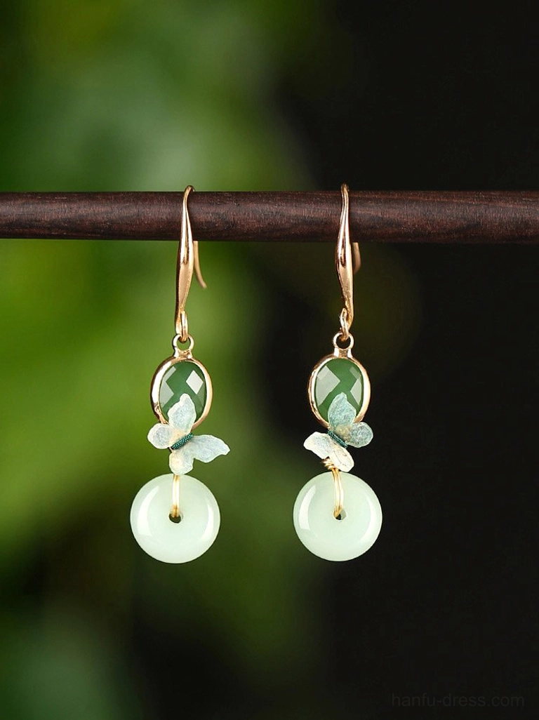 Butterfly jade drop earrings with gold hooks, green oval accents, enamel butterfly charms, and light green peace buckle pendants displayed against a dark botanical background