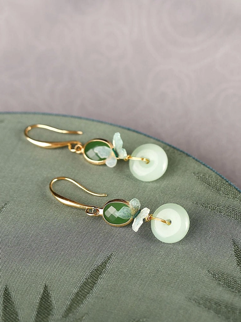 Angled flat lay of butterfly jade earrings with faceted green oval accents, delicate enamel butterflies, gold hooks, and round light green pendants