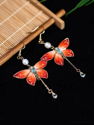 Orange-red butterfly tassel Chinese earrings with gold foil details, velvet flower accents, freshwater pearls, and blue teardrop crystal pendants displayed on a Chinese-style bamboo mat