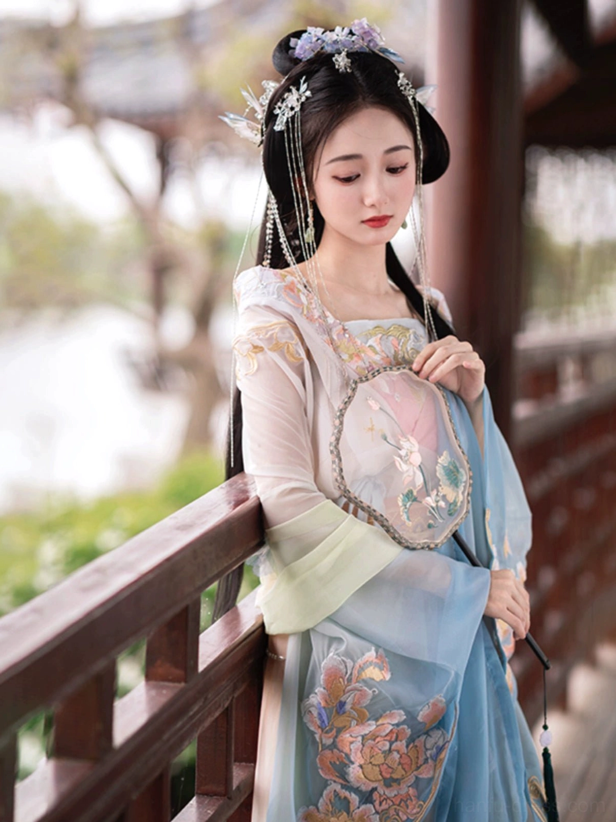 Three-quarter view of Calm Waters Qiyao Ruqun Hanfu beside a traditional wooden lakeside corridor, with peony embroidery, soft blue and apricot colors, and a round silk fan