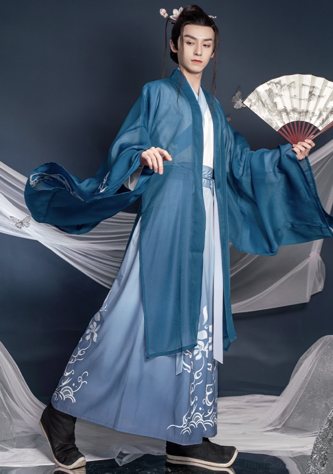 Side full body view of flowing lake blue wuxia hanfu male outfit with embroidered skirt, wide sleeves and folding fan